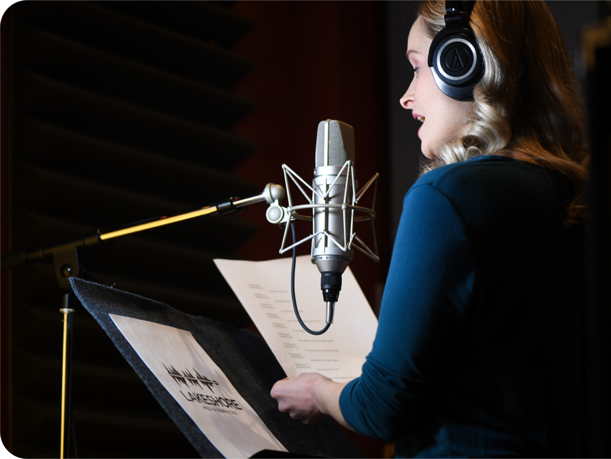 A narrator, a woman with shoulder length blonde hair, wears black headphones and a blue top as she narrates a described video script for Lakeshore Media Services. The narrator stands in a recording booth and speaks into a microphone.